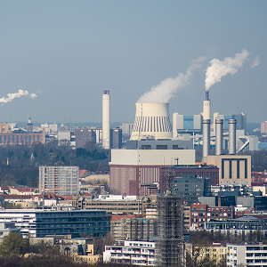 Stadtpanorama mit Industrieanlage/Kraftwerk: großer Kühlturm und mehrere Schornsteine mit Dampf über urbaner Bebauung.